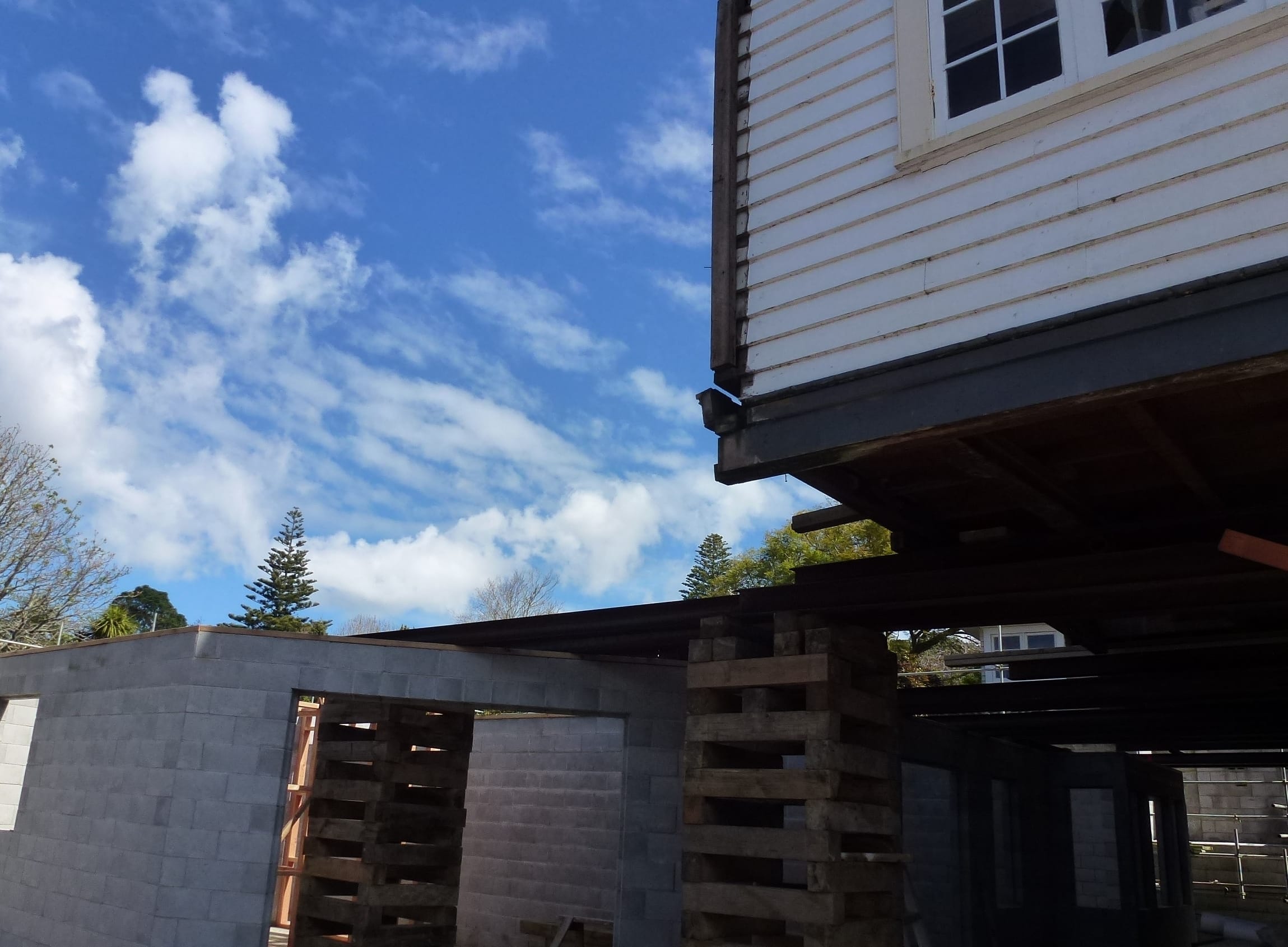 House lifted above newly built concrete block foundation, supported by timber cribbing and steel beams during construction